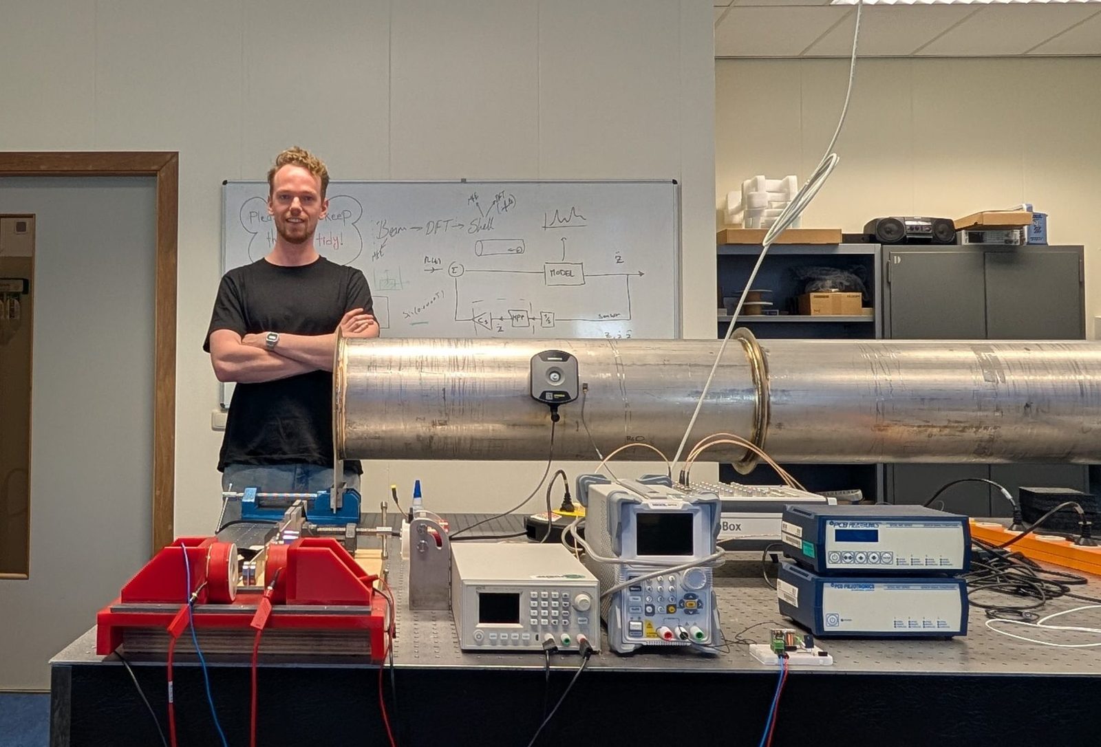 Rinnert Jan Politiek standing behind a steel tube section on a lab table, a block diagram on the whiteboard behind him
