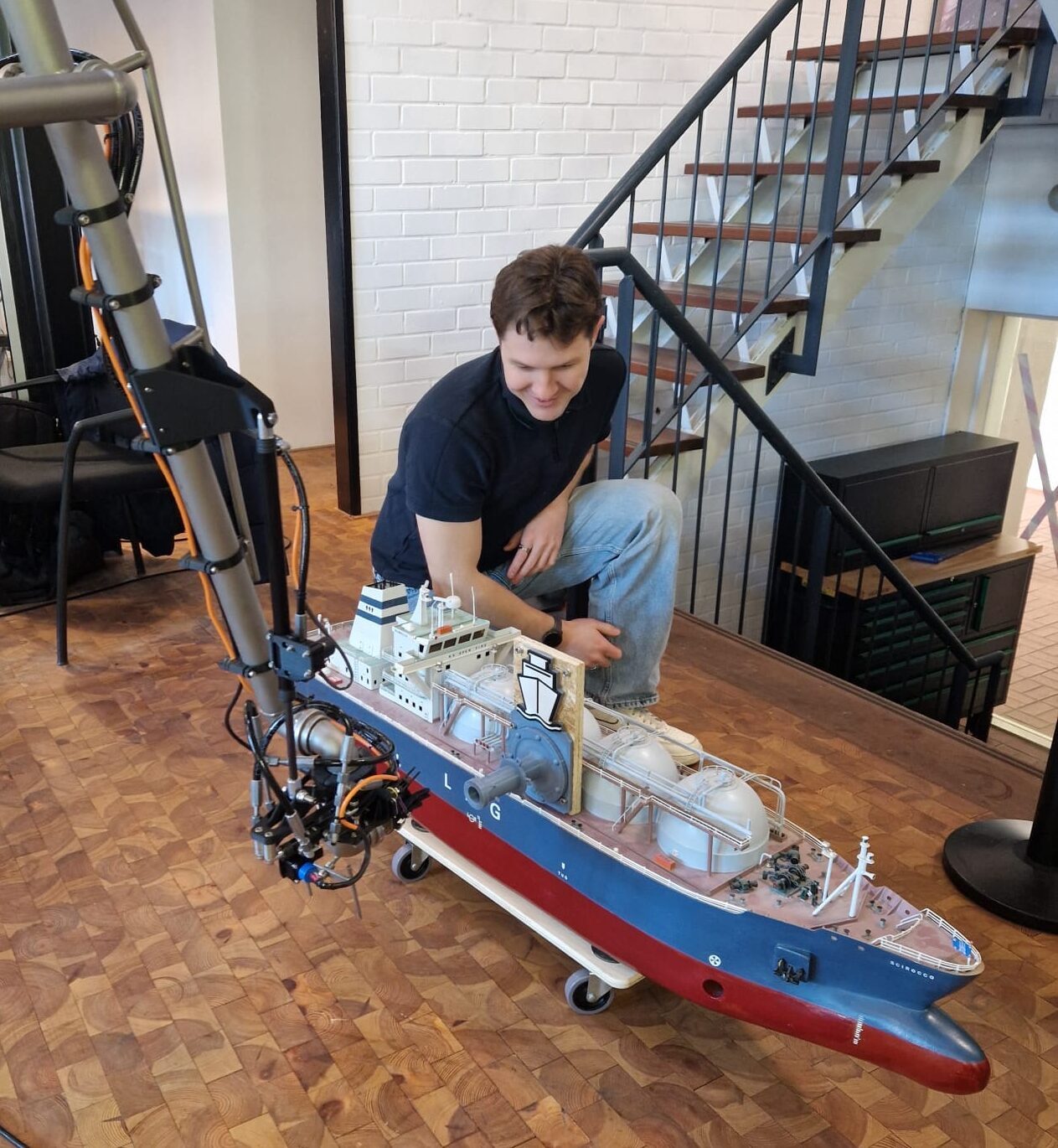 Damien Dufour kneeling beside a scale model of an LNG carrier and a robotic arm