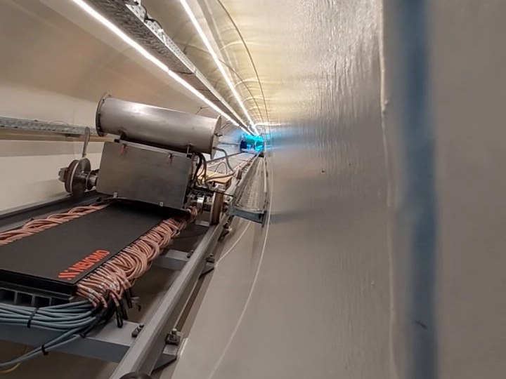 Inside a hyperloop test tube: a pod on its rail at the left, cable trays along the wall and a lit tunnel stretching into the distance