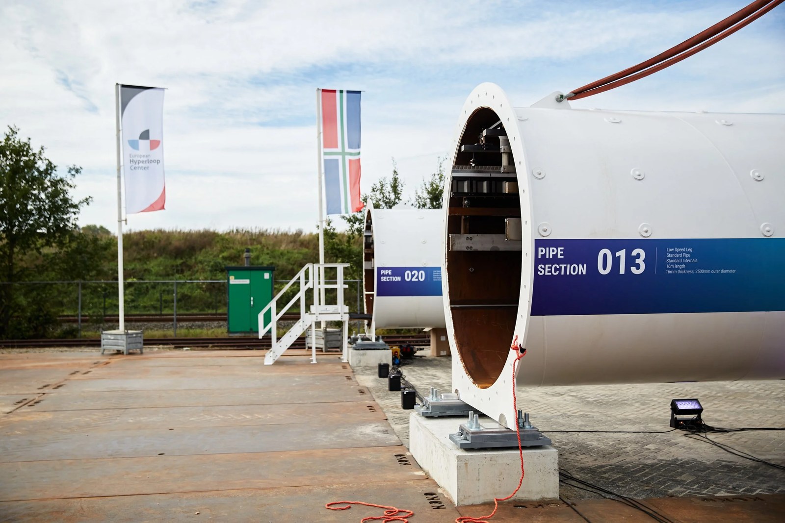 Two large white hyperloop tube sections on concrete supports, labelled pipe section 013 and pipe section 020, with flags of the European Hyperloop Center and the Province of Groningen behind them