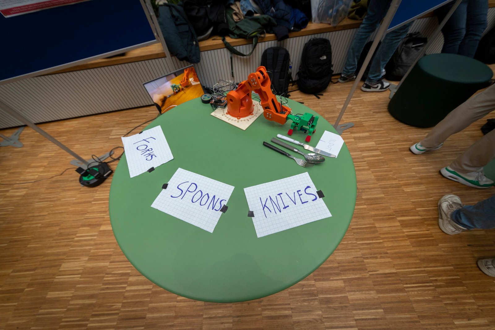An orange printed robot arm with a green gripper on a round green table, next to a knife, a spoon and a fork and three paper labels reading forks, spoons and knives