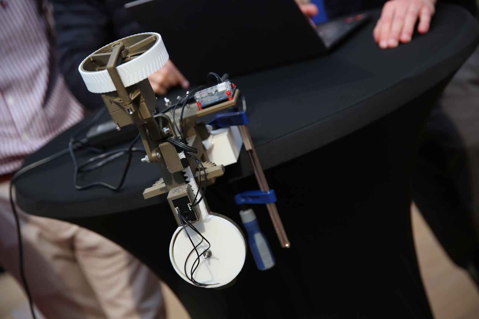 A printed robot arm with a rack-and-pinion extension and a white wheel at its end, fixed to a table edge with two blue bar clamps