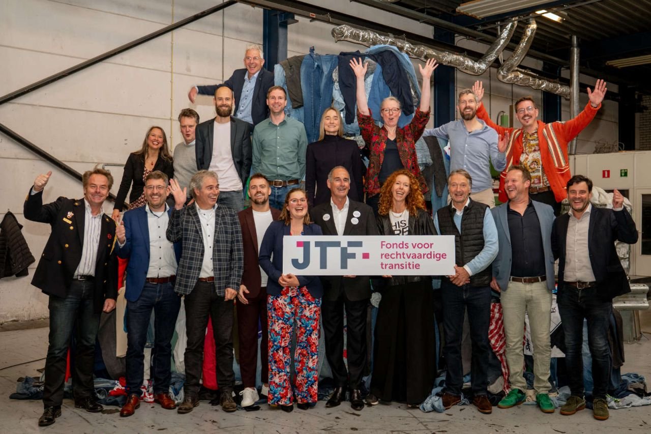 About twenty members of the SORTED consortium in front of a wall of used clothing, holding a Just Transition Fund sign