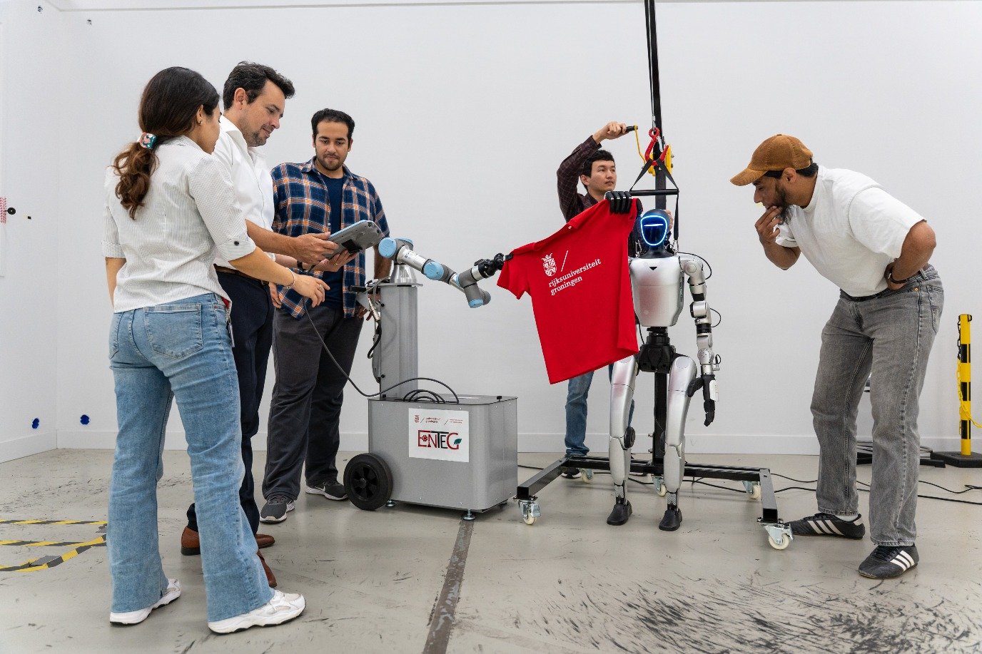Five researchers watch a robot arm and a humanoid robot hold up a red University of Groningen T-shirt together