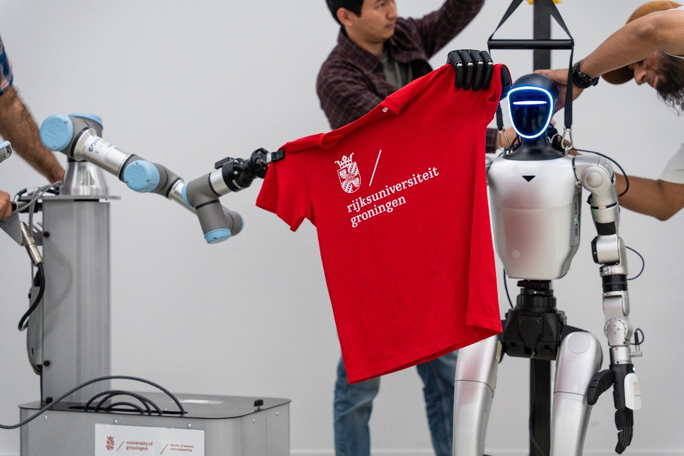 A robot arm grips one shoulder of a red T-shirt while the humanoid holds the other
